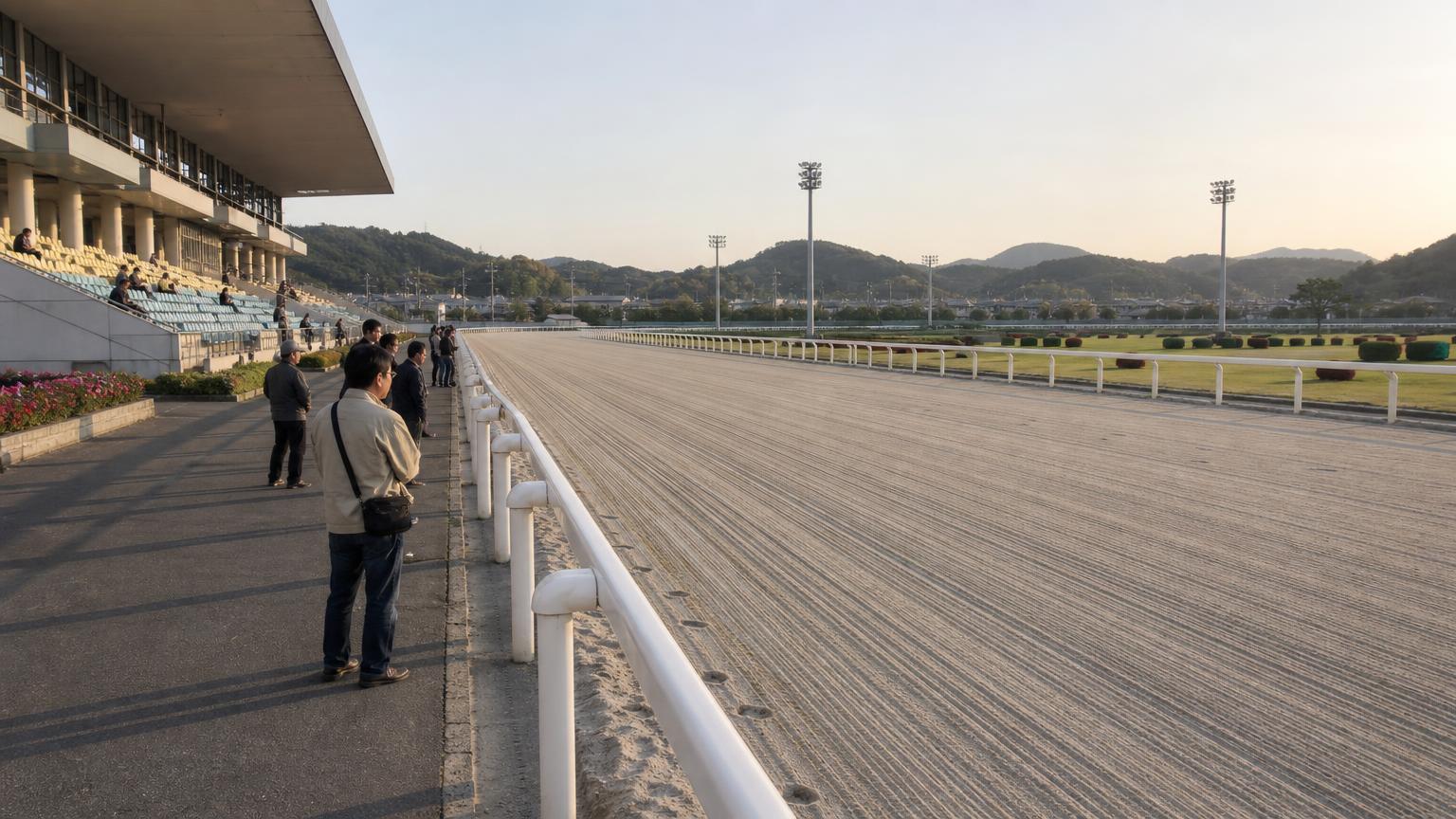 夕方の小さな地方競馬場の全景。手前にスタンドと立ち見の広場、その先の外ラチの向こうに砂のコースが広がり、さらに向こうは内馬場の芝生
