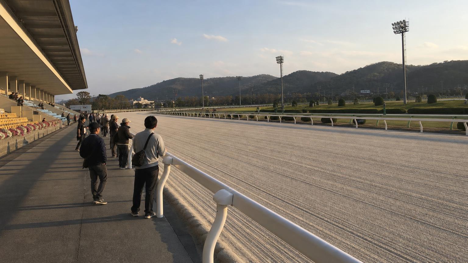 笠松競馬場をイメージした地方競馬場の全景。手前にスタンドと立ち見の広場、その先の外ラチの向こうに砂のコースが広がっている