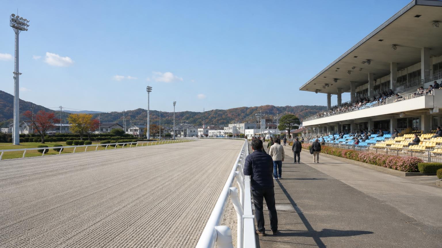 晴れた秋の昼の小さな地方競馬場で、低いスタンドとダートコース、内馬場の向こうの街並みと山並みを見渡した画像